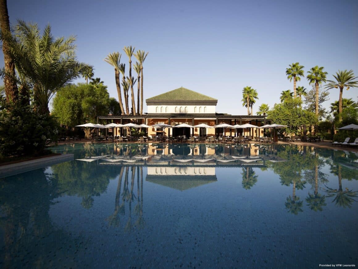 Luxurious exterior view of La Mamounia Marrakesch, featuring a large reflective pool surrounded by lush palm trees and traditional Moroccan architecture with Natlaupa.